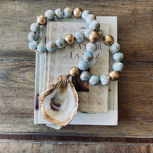 May include: A teal and gold beaded garland with a gold oyster shell hanging from it. The garland is draped over a stack of books with the title "The Prayer of Jabez" visible.
