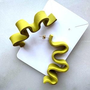 May include: A pair of chartreuse yellow earrings featuring a distinct wavy, ribbon-like design. The earrings are attached to a white rectangular display card, with one earring showing a metallic post back and the other positioned above it.