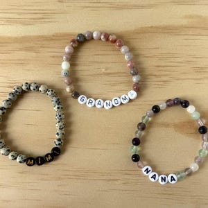 Pode incluir: Três pulseiras de contas dispostas em grupo. Uma pulseira apresenta contas de jaspe dálmata com contas de letras pretas soletrando MIMI. Outra tem contas de pedra multicoloridas com contas de letras brancas soletrando GRANDMA. A terceira tem contas translúcidas multicoloridas com contas de letras brancas soletrando NANA.