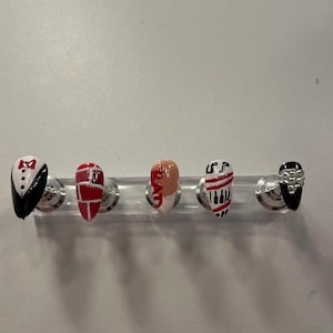 May include: Five almond-shaped press-on nails displayed on a clear stand. The designs include a black tuxedo with a red bow tie, a red and white brick pattern, a nude nail with the word BAD, red and white stripes with music notes, and black with silver studs.
