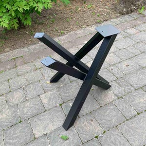 May include: A black metal table base featuring an X-shaped design with four angled legs. The structure is constructed from square tubing and includes flat mounting plates at the top of each leg, each with a single pre-drilled hole for attachment.
