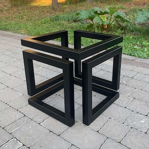 Puede incluir: Una mesa auxiliar negra y geométrica con un diseño de ilusión óptica. La mesa está construida con tubos cuadrados de metal, creando una forma de cubo imposible. La mesa está colocada sobre un patio de piedra gris, con vegetación en el fondo.
