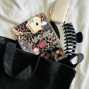 May include: A black tote bag with a leopard print Kindle cover inside. The cover has a collage of images including a disco ball, playing cards, and the word "LUCKY" in silver balloons.