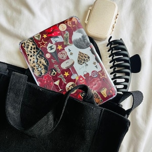 Puede incluir: Una bolsa tote negra con un bolsillo con cremallera y una correa negra. La bolsa está llena de una funda de teléfono transparente con un collage de pegatinas, una pequeña caja beige y una pinza para el pelo negra.