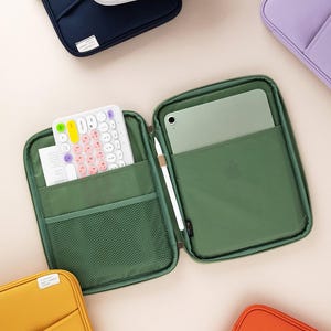 May include: An open green zippered case with a mesh pocket, a white keyboard with colorful keys, and a white stylus in the other pocket. The case is lined with a green fabric and has a zipper closure. The case is open and the keyboard is visible. The case is sitting on a light pink surface.