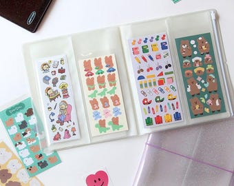 May include: A clear plastic sticker binder with multiple pockets filled with colorful stickers. The stickers feature cartoon bears, school supplies, and other cute designs. There are also some loose stickers on the table, including hearts and a daisy.