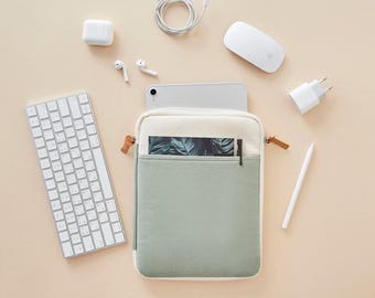 May include: A green and white canvas tablet sleeve with a zipper closure. The sleeve has a pocket on the front for a pen or stylus. The sleeve is lying flat on a light beige surface.