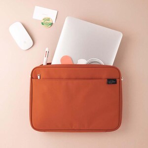 May include: A burnt orange laptop sleeve with a zipper closure and a small pocket on the front. The sleeve is made of a durable fabric and has a black label with the text "A Line Made" on it.