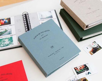 May include: A blue, green, and beige photo album with a ring binder. The album is labeled "Album de Polaroid pour Ma Memoire" and has a number "120" printed on the front. The album is on a white surface with several Polaroid photos.