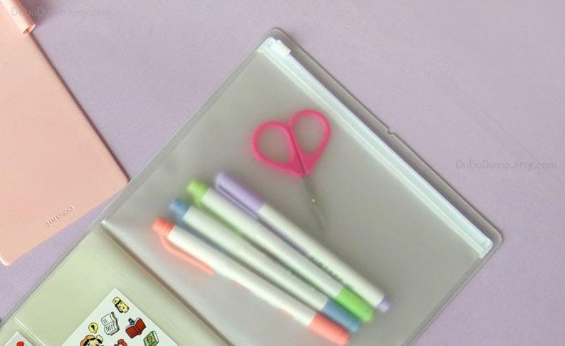 Może przedstawiać: Przezroczysty plastikowy woreczek z zamkiem błyskawicznym, w kt&oacute;rym znajdują się r&oacute;żowe nożyczki w kształcie serca i pięć kolorowych długopis&oacute;w. Woreczek leży na jasnofioletowym tle.