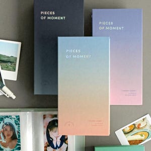 May include: Three photo albums with the text "Pieces of Moment" on the front. The albums are in different colors: black, pink and blue. The albums are on a gray surface.