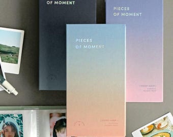 May include: Three photo albums with the text "Pieces of Moment" on the front. The albums are in different colors: black, pink and blue. The albums are on a gray surface.