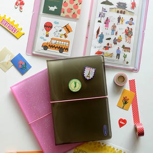 May include: A pink and brown plastic binder with a clear plastic pocket on the front. The pocket contains colorful stickers and illustrations. The binder is open and there are other stickers and washi tape on the table.