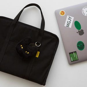 May include: A black canvas laptop bag with a zipper closure and two top handles. A black plush cat keychain with gold accents is attached to the bag.