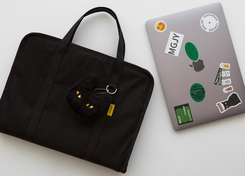 May include: A black canvas laptop bag with a zipper closure and two top handles. A black plush cat keychain with gold accents is attached to the bag.