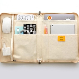May include: A beige canvas laptop bag with multiple pockets and compartments. The bag is open and filled with various items, including a laptop, a tablet, a mouse, a notebook, pens, and a power adapter.