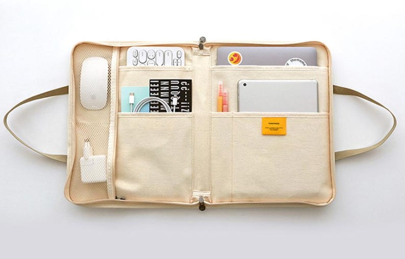 May include: A beige canvas laptop bag with multiple pockets and compartments. The bag is open and filled with various items, including a laptop, a tablet, a mouse, a notebook, pens, and a power adapter.