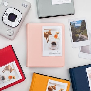 May include: Four photo albums in different colors: red, pink, yellow, and blue. Each album has a clear plastic window to display a photo. The text on the pink album reads "instax album easy square wide".