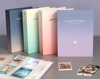 May include: Four photo card albums with ombre covers in blue, green, pink, and purple. The albums are labeled "Pieces of Moment" and have a gradient design. The albums are on a gray surface with some scattered glitter.