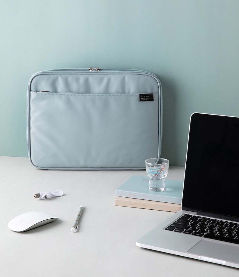 May include: A light blue laptop sleeve with a zip fastening and a small pocket on the front. The sleeve is sitting on a white surface next to a laptop, a glass, and a stack of notebooks.