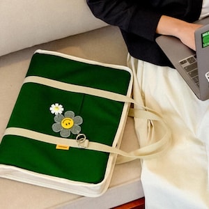 May include: A green canvas tote bag with a white trim and a smiling flower patch. The bag has a long strap and is sitting on a white surface.