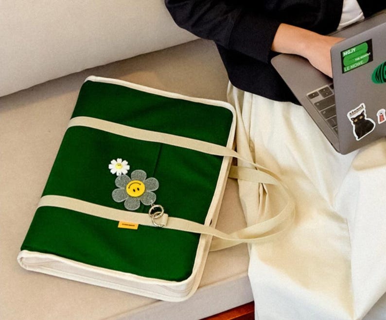 May include: A green canvas tote bag with a white trim and a smiling flower patch. The bag has a long strap and is sitting on a white surface.