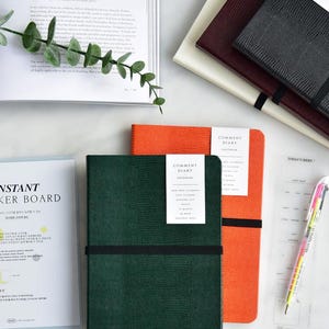 May include: Three green, orange, and black faux leather journals with the text "Comment Diary" on the front cover. The journals are stacked on top of each other.