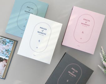 May include: Four photo albums with different colored covers. The covers are light blue, white, pink, and gray. Each cover has a circle with the text "Pieces of Moment" in a navy blue font.