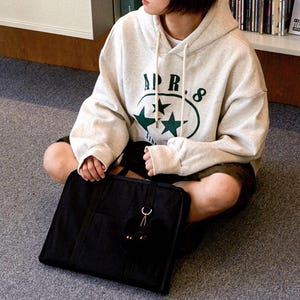 May include: A person wearing a cream colored hoodie with the text "APR. 8" and two green stars in a circle. The person is holding a black tote bag with a black fuzzy keychain.