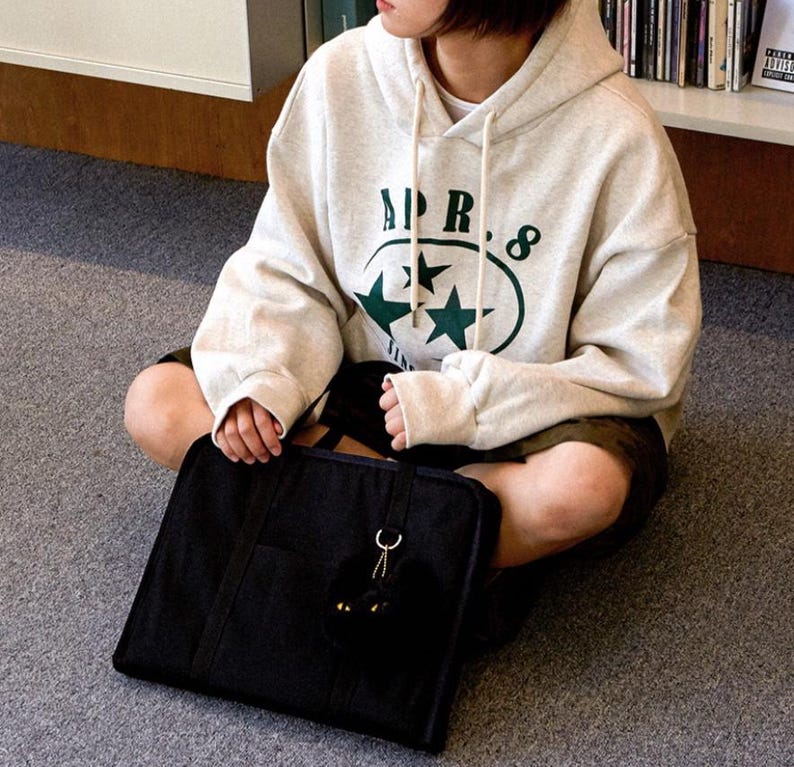 May include: A person wearing a cream colored hoodie with the text "APR. 8" and two green stars in a circle. The person is holding a black tote bag with a black fuzzy keychain.