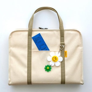 May include: A beige canvas tote bag with a zipper closure and two long straps. The bag has a front pocket with a blue ticket inside and a white daisy keychain with a yellow smiley face.
