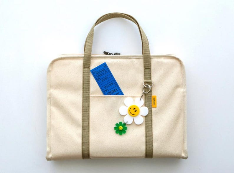May include: A beige canvas tote bag with a zipper closure and two long straps. The bag has a front pocket with a blue ticket inside and a white daisy keychain with a yellow smiley face.