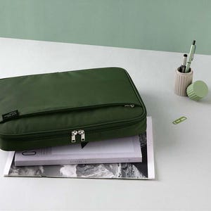 May include: A dark green zippered laptop case with a small pocket on the front. The case is sitting on top of a magazine.