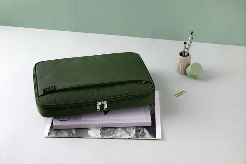 May include: A dark green zippered laptop case with a small pocket on the front. The case is sitting on top of a magazine.