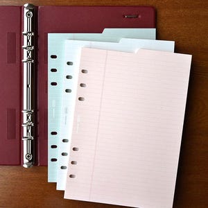 May include: A red binder with three different colored paper refills. The refills are lined and have holes for a binder. The paper is light blue, white, and pink.