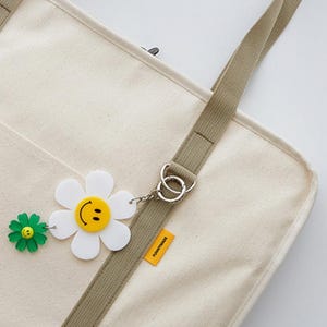 May include: A white canvas tote bag with a brown strap and a daisy charm with a smiley face. The bag has a pocket on the front and a small tag that says "Tomorrowland".