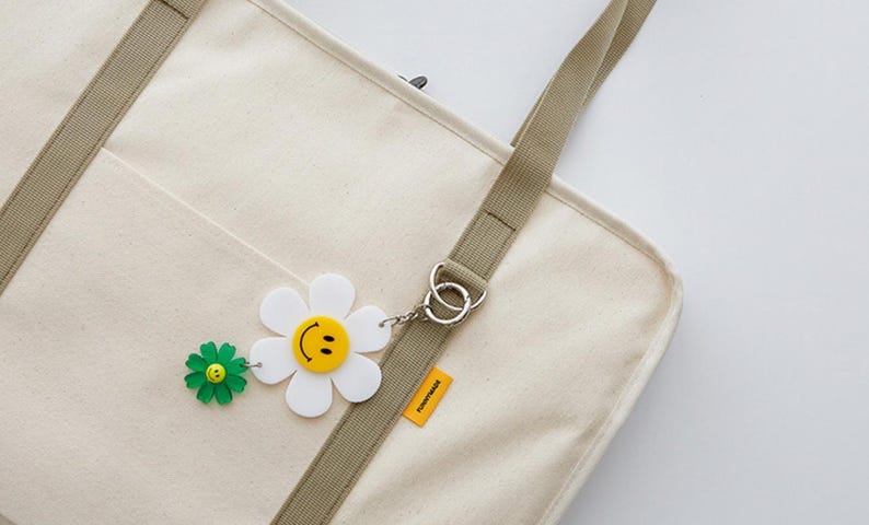 May include: A white canvas tote bag with a brown strap and a daisy charm with a smiley face. The bag has a pocket on the front and a small tag that says "Tomorrowland".