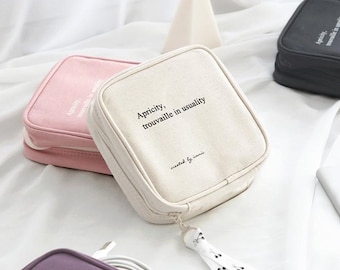 May include: Three zippered pouches in pink, beige, and purple with the text "Apricity, trouvaille in usuality" and "created by iconic" on the beige pouch. The pouches have a zipper closure and a small tassel with a cherry design.