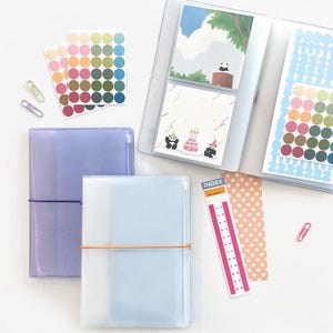 May include: Two clear plastic binders with elastic closures. The binder on the left is lavender and the binder on the right is light blue. Both binders are closed and have a few pages inside. There are also several sheets of colourful stickers and a pink index card with the word "INDEX" on it.