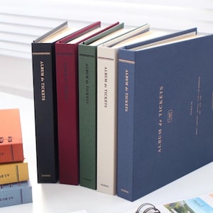 May include: Six different colored photo albums with the text "ALBUM de TICKETS" on the spine. The albums are arranged in a row from left to right, with the black album on the left and the blue album on the right.