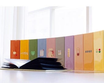 May include: A set of 12 colorful photo albums with various cute illustrations and text. The albums are arranged in a semi-circle on a white surface. The albums are all the same size and shape, and they are all closed.