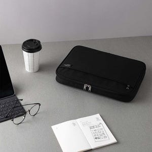 May include: Black zippered pouch with a silver zipper pull. The pouch is on a grey surface with a coffee cup, a laptop, glasses, and a notebook.