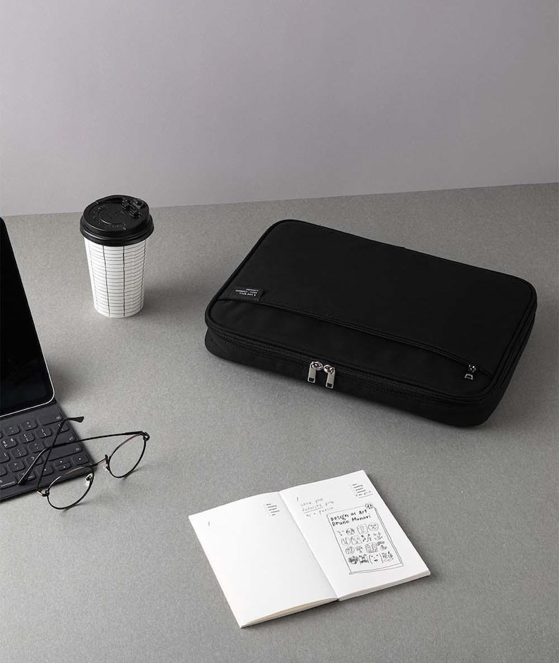 May include: Black zippered pouch with a silver zipper pull. The pouch is on a grey surface with a coffee cup, a laptop, glasses, and a notebook.