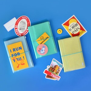 May include: Three clear plastic notebooks with colorful stickers and a yellow rubber band. The notebooks are on a blue background. The stickers include a bunny, a sad face, a smiley face, a duck, and a building with the text "Hotel Sudan".