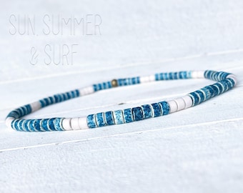 May include: A blue and white beaded necklace with a gold clasp. The beads are small and round, and they are arranged in a pattern of blue and white. The necklace is on a white wooden surface.