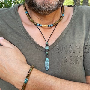 Op de afbeelding: Een man draagt een groen T-shirt met de tekst "SUN. SUMMER & SURF" erop gedrukt. Hij draagt een gelaagde ketting met een houten kralenstreng en een leren koord met een turquoise hanger. Hij draagt ook een houten kralenarmband om zijn pols.