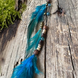 May include: A blue feather hair tie with wooden and turquoise beads. The hair tie has the text "SUN. SUMMER & SURF" on a wooden background.