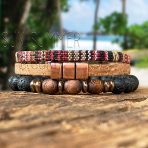 May include: Three layered bracelets featuring a woven fabric band, a cork band, and a beaded band with copper accents. The bracelets are stacked on a wooden surface.