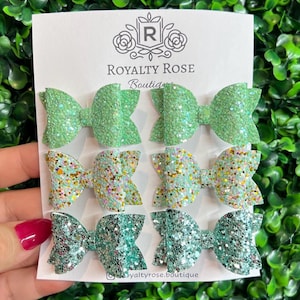 May include: Six hair bows arranged in two columns on a white display card labeled Royalty Rose Boutique. The top bows are light green glitter, the middle bows are mint green with gold and white glitter, and the bottom bows are teal glitter.