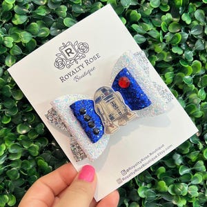 May include: A hair bow featuring white and silver glitter fabric, accented with blue glitter sections, a central R2-D2 character graphic, a red rhinestone, and black studs. The bow is attached to a Royalty Rose Boutique display card.